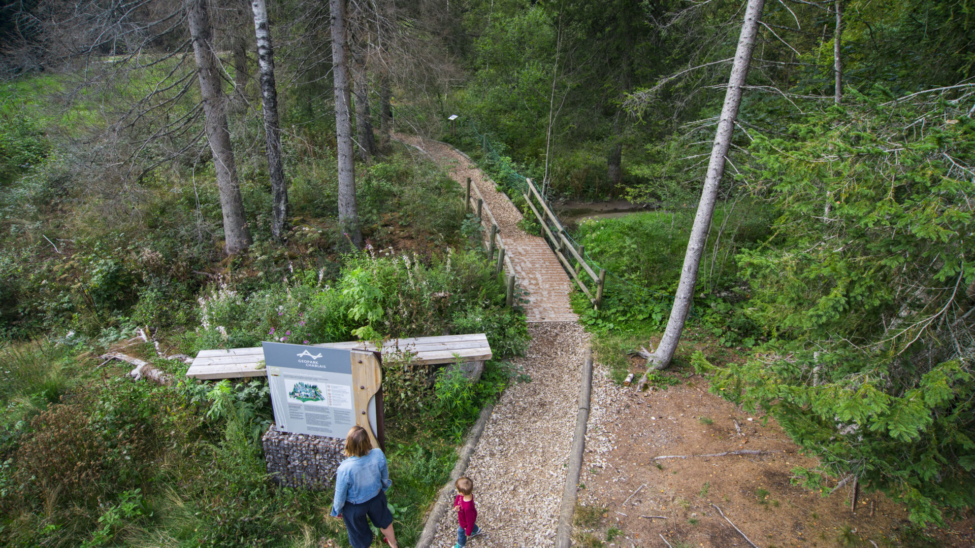 Femme et son enfant regardant un panneau explicatif Géopark Chablais. Chemin de promenade en forêt